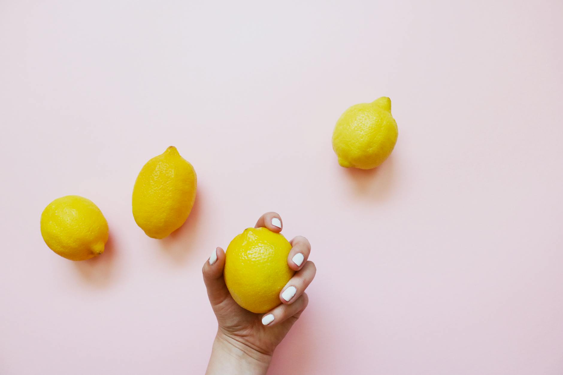 A hand with white nails holding a bright lemon on a soft pink background